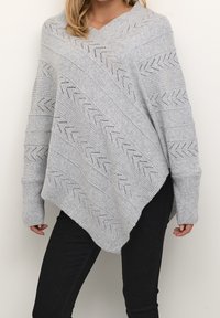 Kvinna klädd i en lös, ljusgrå stickad poncho med chevronmönster, kombinerad med svarta byxor, som står mot en enkel ljus bakgrund.