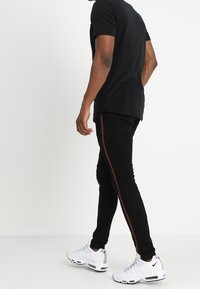 Jeans negros ajustados con una franja lateral roja, combinados con una camiseta negra y zapatillas deportivas blancas. Textura suave, silueta esbelta.