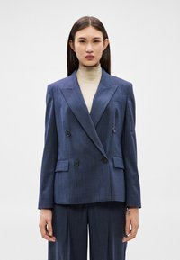 Žena v tmavě modrém dvouřadém saku a kalhotách v odpovídající barvě s béžovým rolákem, stojící před jednobarevným bílým pozadím.