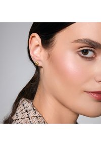 Złoty, sferyczny kolczyk wkręcany, z polerowanym wykończeniem. Zbliżenie pokazuje anatomię ucha i drobne detale twarzy. Widoczny teksturowany, jasny materiał kołnierza.