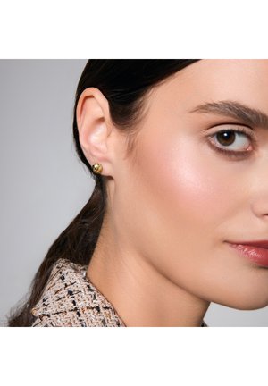 Złoty, sferyczny kolczyk wkręcany, z polerowanym wykończeniem. Zbliżenie pokazuje anatomię ucha i drobne detale twarzy. Widoczny teksturowany, jasny materiał kołnierza.