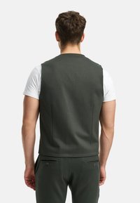 Dunkelgrüne ärmellose Weste aus strukturiertem Stoff, mit schmalem Schnitt und ohne sichtbare Hardware, getragen über einem weißen T-Shirt.
