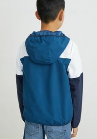 Dětská barevně blokovaná bunda s designem v modré, bílé a námořnické barvě, s kapucí a elastickými manžetami. Vyrobena z lehkého, hladkého materiálu.