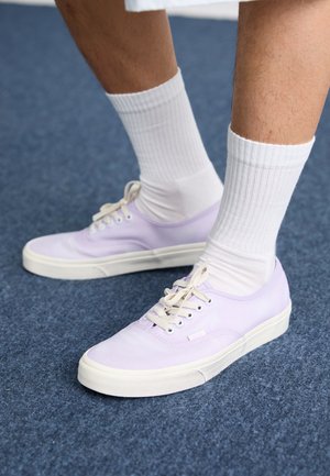 Lila Leinenschuhe mit weißen Gummisohlen und Schnürsenkeln, kombiniert mit weißen gerippten Socken, auf einer blauen Filzoberfläche.