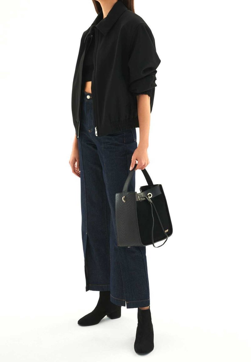 Femme portant une veste courte noire, un haut noir, un jean large foncé, des bottines noires, tenant un sac à main noir avec deux poignées.