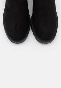 Botines de ante negro con punta redondeada y suela de goma plana, que presentan un detalle de borde texturizado para mayor agarre y estilo.