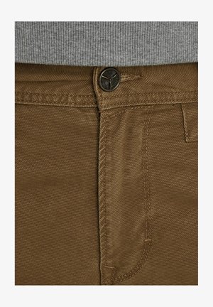 Brune bomuldsbukser med en struktureret vævning, en metalsknap og kontrastsyninger langs bindebåndet og lommerne.