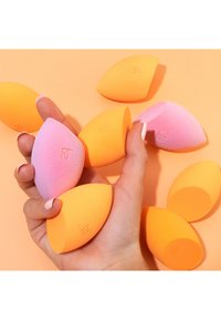 Hand holding several pink and orange makeup blending sponges against an orange background.