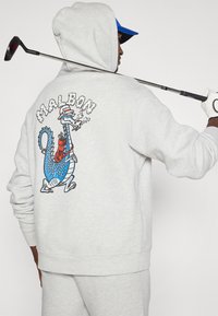 Γκρι φούτερ με σχέδιο πολύχρωμου καρτούν δράκου στην πλάτη, που κρατάει ένα ποτό, με κουκούλα και ραμμένες μανσέτες. Ορατό το γκολφ σάιτ.