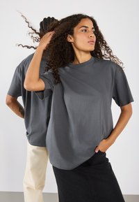 Grå kortærmet t-shirt med en afslappet pasform og rund halsudskæring, parret med en sort nederdel, præsenteret mod en neutral baggrund.