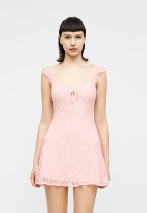 Ung kvinne med kort mørkt hår iført en ermeløs rosa blonderkjole med en liten sløyfedetalj ved halslinningen, stående foran en ensfarget hvit bakgrunn.