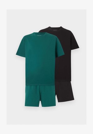 Férfi fekete túlméretezett pólóban, fekete rövidnadrágban, fekete sportcipőben, fehér zokniban és napszemüvegben, egy egyszínű fehér háttér előtt áll.