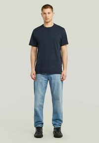 T-shirt blu navy a maniche corte con scollatura rotonda, abbinato a jeans blu chiaro e sneakers nere. Presenta un piccolo logo sul petto.