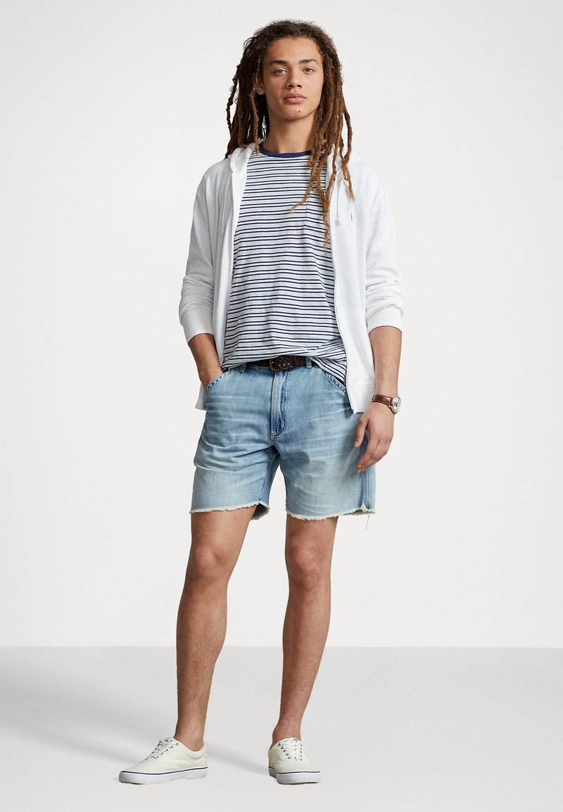 Weiße Zip-Up-Kapuze über einem navy- und weiß gestreiften Hemd, kombiniert mit hellblauen Denim-Shorts und weißen Sneakern. Schlichter brauner Gürtel, lässiger Look.