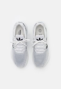 Pár bílých a šedých tenisek Adidas s černými logy a bílými tkaničkami, zobrazený z vrchního pohledu na bílém pozadí.