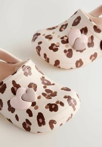 Sabots en imprimé léopard beige clair avec des taches brunes, dotés de deux boutons en forme de cœur rose sur le côté et de trous de ventilation.