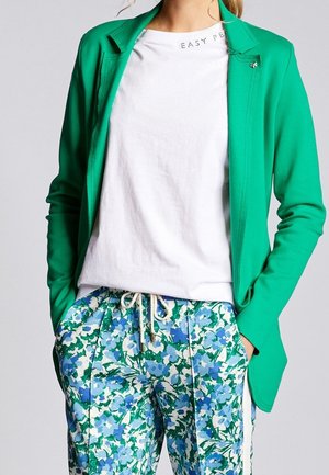 Frau trägt ein weißes T-Shirt, einen leuchtend grünen Blazer und blau-grüne, geblümte Hose mit Kordelzug, Hände in den Taschen.