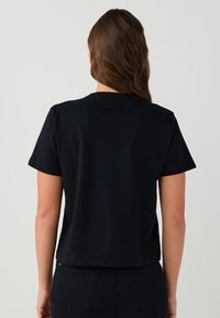 Vrouw met lang bruin haar draagt een eenvoudige zwarte t-shirt met korte mouwen en zwarte broek, gefotografeerd van achteren tegen een effen lichte achtergrond.