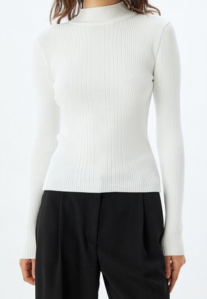 Femme portant un pull à col roulé blanc côtelé ajusté à manches longues et un pantalon noir ample, vue du cou jusqu'aux hanches.