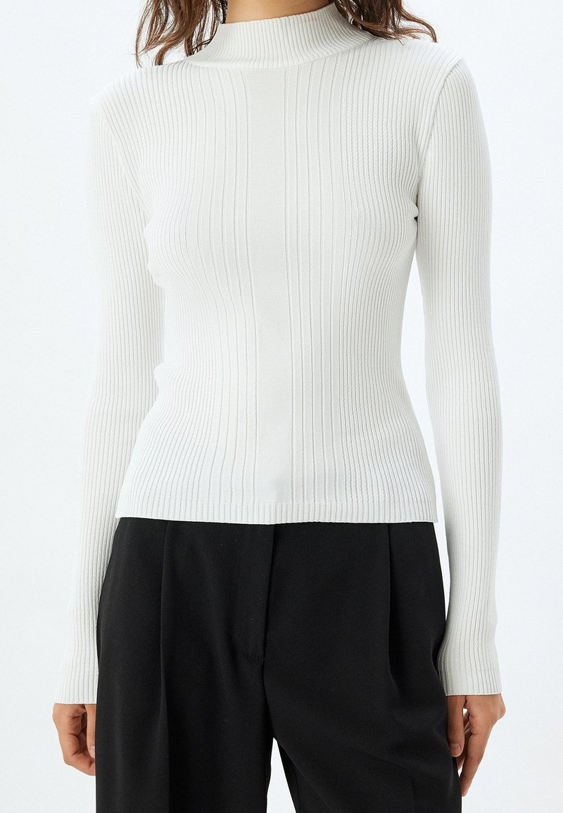 Femme portant un pull à col roulé blanc côtelé ajusté à manches longues et un pantalon noir ample, vue du cou jusqu'aux hanches.