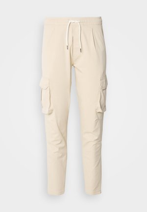Beżowe spodnie cargo z elastycznym pasem i sznurkiem. Posiadają dwie boczne kieszenie cargo oraz zwężający się krój nogawek. Gładka faktura.