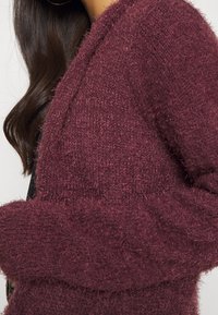 Cardigan lavorato a maglia bordeaux con una texture morbida, apertura frontale e maniche larghe. Il materiale sembra morbido e caldo, adatto per essere indossato a strati.