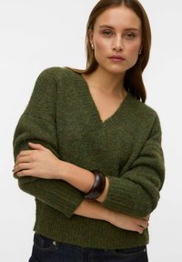 Grüner Strickpullover mit V-Ausschnitt, gerippten Bündchen und Saum. Weiche Textur mit subtilen Farbvariationen, kombiniert mit einem dunklen Armband.
