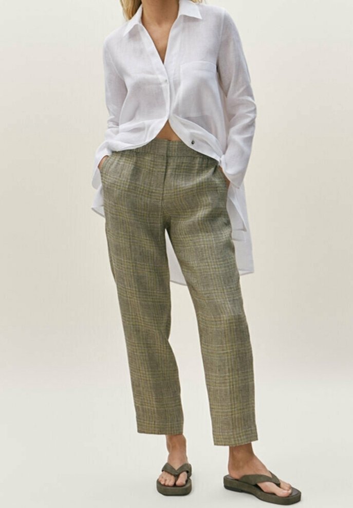 Femme portant une chemise blanche à manches longues boutonnée, un pantalon taille haute à carreaux vert olive et des sandales tongs vert foncé, debout les mains dans les poches.