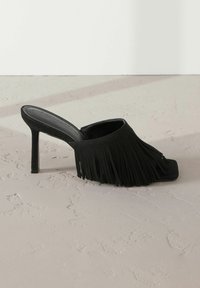 Mules à talons noirs avec des détails frangés, bout carré et dessus en suède texturé. Talon aiguille haut et design ouvert à l'arrière. Semelle plate.