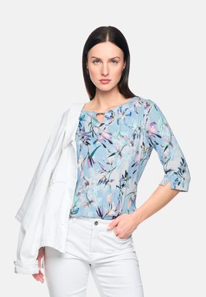 Woman wearing a light blue floral blouse with three-quarter sleeves, white pants, and a white jacket draped over her shoulder.