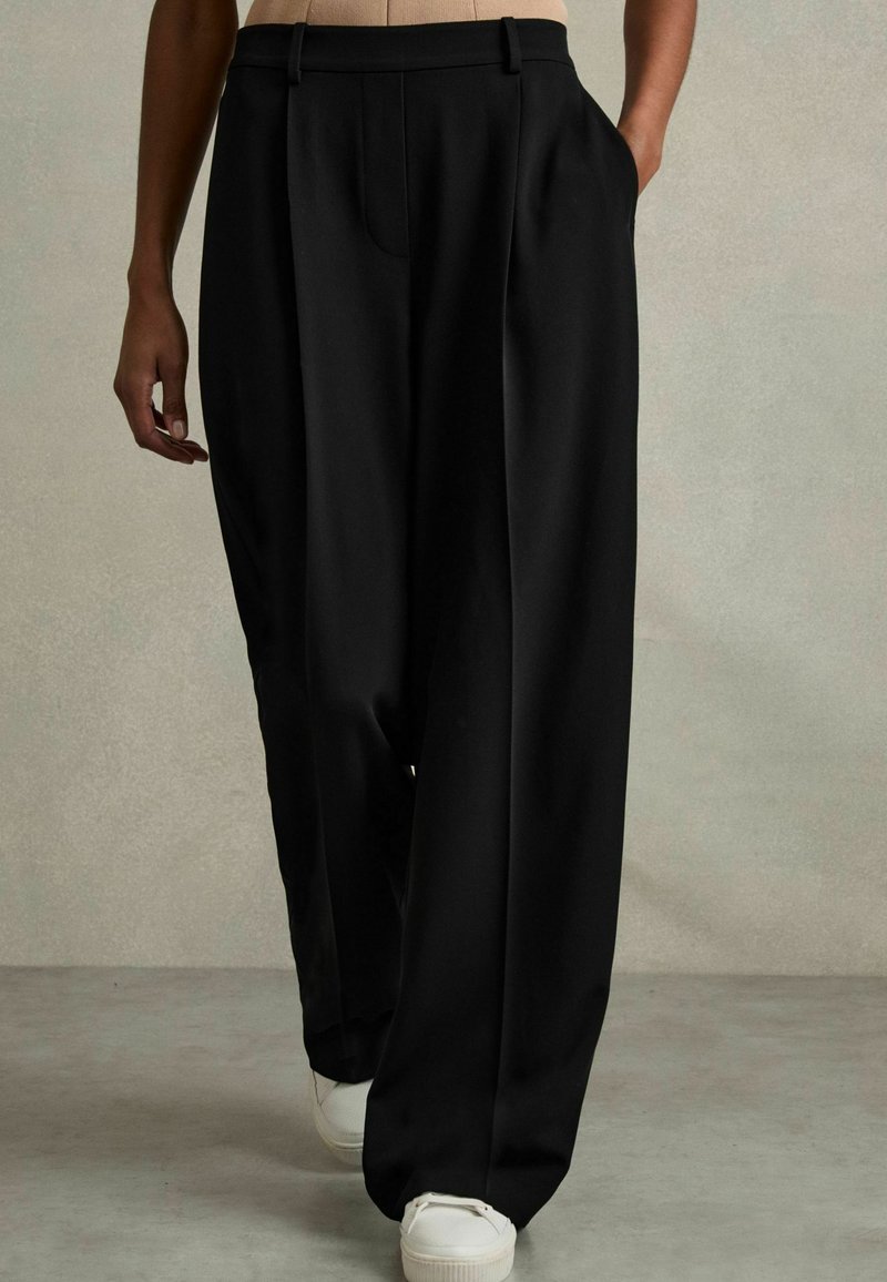 Pantalons larges noirs avec une texture lisse, dotés d'un pli avant et de deux poches latérales, associés à des baskets blanches.