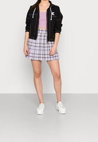 Giacca nera con zip e dettagli bianchi, top corto viola chiaro e gonna plissettata a quadri in viola e giallo. Sneakers bianche completano l'outfit.