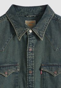 Σκούρο μπλε πουκάμισο από τζιν με μυτερό γιακά, κουμπιά κλικ, τσέπη στο στήθος και αντίθετες ραφές. Ο ετικέτα με το λογότυπο της Levi's είναι ορατή στο πλάι.