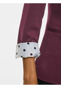 Bras portant une veste bordeaux avec le revers relevé pour révéler un tissu blanc à pois noirs.