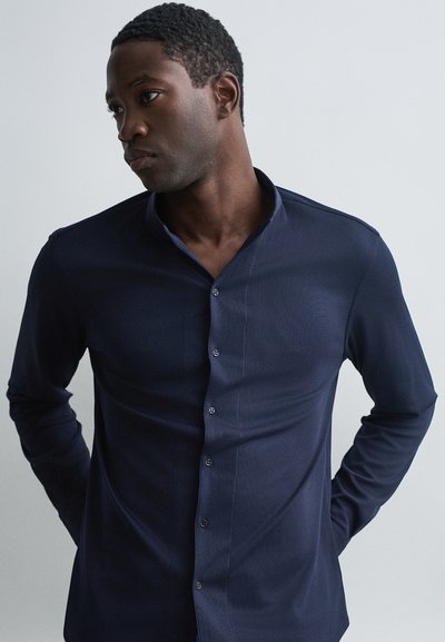 Chandail bleu marine à manches longues avec un col structuré, une texture à côtes verticales et des boutons sur le devant ; la coupe ajustée met en valeur la silhouette.