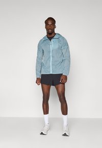 Lichtblauwe lichte jas met capuchon, voorkleurige rits en borstzak, gecombineerd met zwarte shorts en grijze sportschoenen.