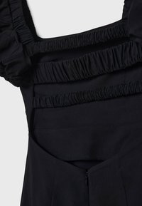 Robe en tissu noir avec bretelles élastiques froncées sur un dos ouvert et une fermeture éclair visible au centre inférieur.