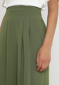 Naisen oikea käsi lepäilee sivulla, hänellä on päällään oliivivihreä laskostettu hame ja valkoinen paita yksinkertaista taustaa vasten.