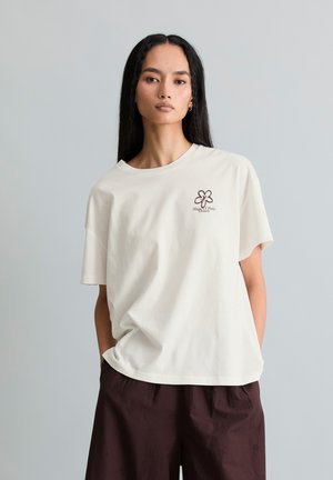 Frau mit langen schwarzen Haaren trägt ein lockeres cremefarbenes T-Shirt mit kleinem Blumenlogo und dunkelbraune lockere Hose, steht vor einfachem Hintergrund.