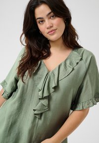 Blusa verde a volant con scollo a V e maniche corte, realizzata in un tessuto texturizzato e con una vestibilità ampia. I volant accentuano lo scollo.