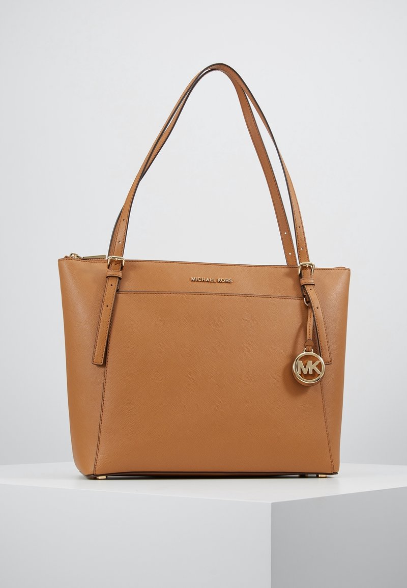 Sac fourre-tout en cuir tan de Michael Kors avec deux anses réglables, un charm logo en or et une poche avant sur un piédestal blanc.