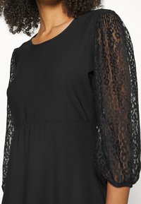Vestido negro con un cuerpo entallado, que presenta mangas de volante de encaje transparente en un patrón de leopardo. Textura suave con cuello redondo.
