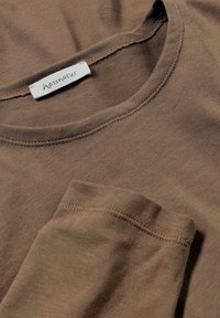 Chemise marron à manches longues avec un col rond et une texture en coton doux. Présente une étiquette hessnatur près du col et un ourlet soigneusement cousu.