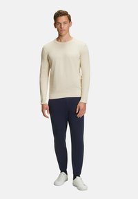 Camicia a maniche lunghe crema con collo rotondo, abbinata a pantaloni aderenti blu e sneakers bianche. Tessuto liscio con un design semplice.