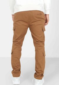 Bruine cargobroek met taps toelopende benen, voorzien van zijzakken en elastische boorden. De stof lijkt stevig en glad, in combinatie met witte sneakers.