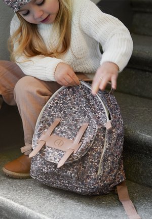 Junges Mädchen in weißem Pullover und Stiefeln öffnet einen Rucksack mit Blumenmuster, während sie auf einer Innentreppe sitzt.