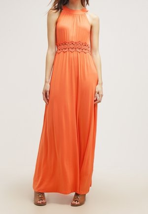 Vrouw die een oranje mouwloze jurk draagt die tot de vloer reikt, met kantdetail bij de taille en een geplooide bovenkant, gecombineerd met beige open-toe hakken.