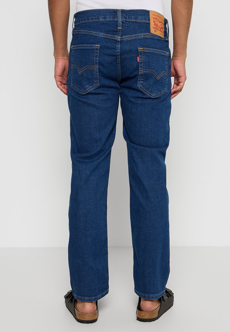 Mann in dunkelblauen, geraden Levi's Jeans, der mit dem Rücken steht, mit braunen Sandalen und einem weißen Oberteil.