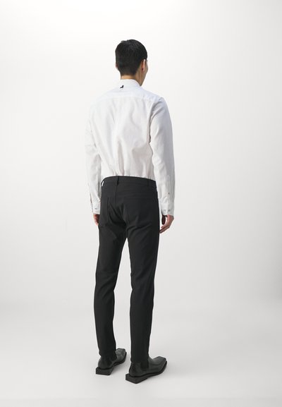 Chemise blanche à manches longues avec un empiècement au dos, associée à un pantalon noir slim et des chaussures noires à enfiler, vue de dos.