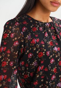 Blusa nera con stampa floreale caratterizzata da un mix di fiori rossi e viola, con una consistenza morbida e trasparente e un dettaglio di scollatura arricciata.